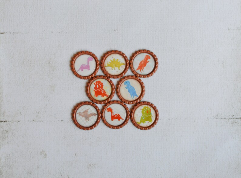 May include: Nine brown round bottle caps with different dinosaur designs. The dinosaurs are in various colors, including pink, orange, blue, green, and yellow.