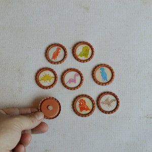 May include: Eight round, brown, paper-like stickers with different dinosaur designs. The stickers have a white background and feature colorful dinosaurs. One sticker is a plain brown circle.
