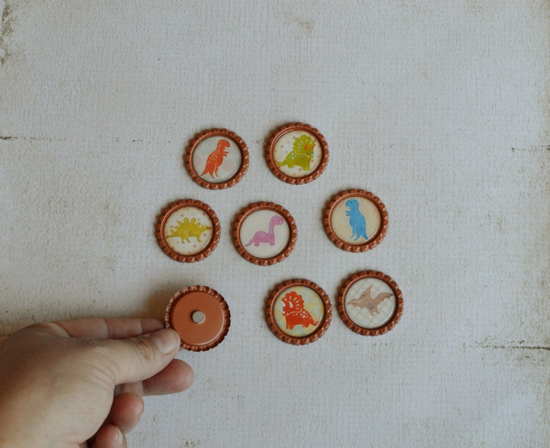 May include: Eight round, brown, paper-like stickers with different dinosaur designs. The stickers have a white background and feature colorful dinosaurs. One sticker is a plain brown circle.