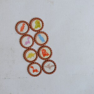 May include: Eight round bottle caps with different dinosaur designs. The caps are brown with a gold rim and feature colorful dinosaurs on a white background.