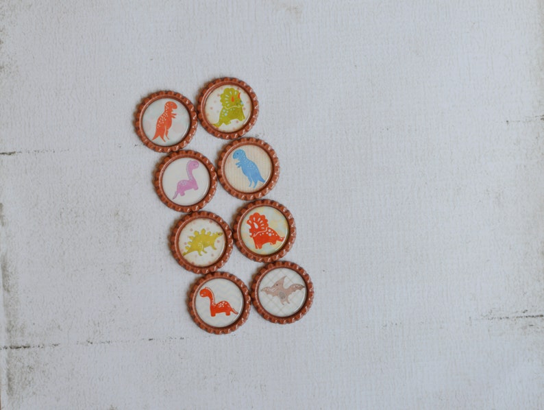 May include: Eight round bottle caps with different dinosaur designs. The caps are brown with a gold rim and feature colorful dinosaurs on a white background.