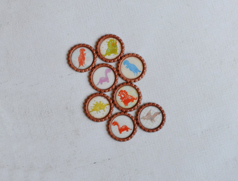 May include: Eight brown round bottle caps with different dinosaur designs. The dinosaurs are in various colors, including red, blue, green, and purple.