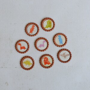 May include: Eight round brown bottle caps with different dinosaur designs. Each cap features a different dinosaur in various colors, including orange, pink, blue, and green.