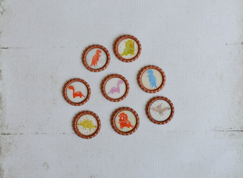 May include: Eight round brown bottle caps with different dinosaur designs. Each cap features a different dinosaur in various colors, including orange, pink, blue, and green.