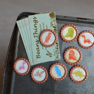 May include: Set of eight round magnets with a copper-colored metal frame and a clear plastic top. Each magnet features a different dinosaur design with colorful illustrations. The magnets are arranged on a metal surface with a business card that reads "Beans Things" with a website address.