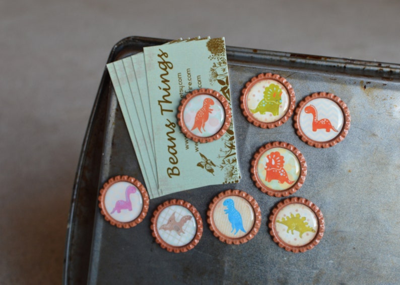 May include: Set of eight round magnets with a copper-colored metal frame and a clear plastic top. Each magnet features a different dinosaur design with colorful illustrations. The magnets are arranged on a metal surface with a business card that reads "Beans Things" with a website address.