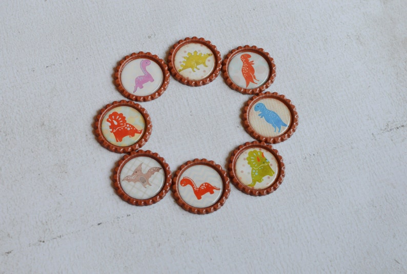 May include: Eight brown bottle caps with different dinosaur designs. Each cap features a different dinosaur in a colorful cartoon style.
