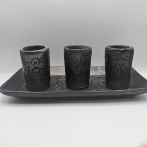Può includere: Tre portacandele neri in ceramica con motivi floreali, posti su un vassoio rettangolare abbinato. I portacandele cilindrici e il vassoio presentano una superficie strutturata, creando un insieme armonioso.