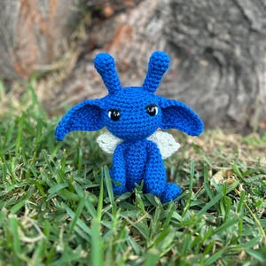 Puede incluir: Una criatura de crochet azul con orejas grandes y alas blancas. La criatura está sentada en un lecho de hierba verde.