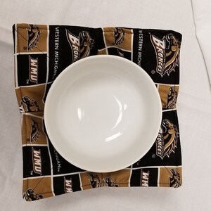 Op de afbeelding: Een zwarte en gouden stoffen kommetjeswarmer met het logo van de Western Michigan University Broncos. De kommetjeswarmer is vierkant en gemaakt van een zachte, gestikte stof. Het logo is een bruin paardenhoofd met de woorden "Western Michigan" en "Broncos" eronder.