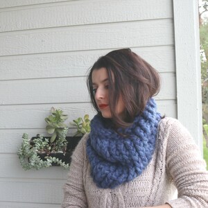May include: A woman wearing a blue knitted infinity scarf. The scarf is thick and chunky, and the woman is wearing a beige sweater.