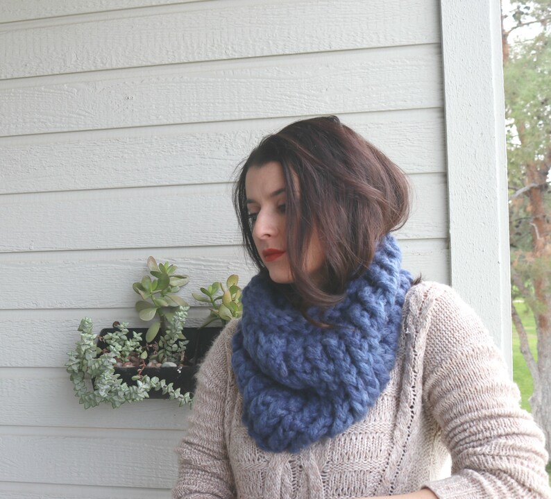 May include: A woman wearing a blue knitted infinity scarf. The scarf is thick and chunky, and the woman is wearing a beige sweater.