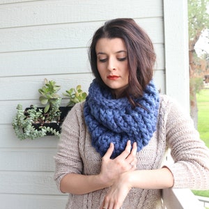 May include: A blue knitted infinity scarf with a chunky knit texture. The scarf is worn by a person with long brown hair.