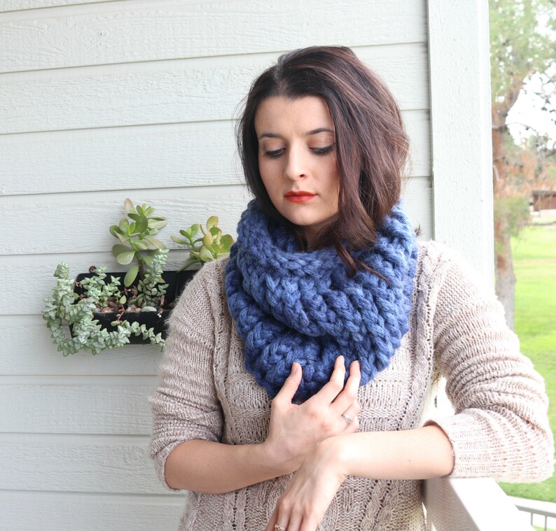 May include: A blue knitted infinity scarf with a chunky knit texture. The scarf is worn by a person with long brown hair.