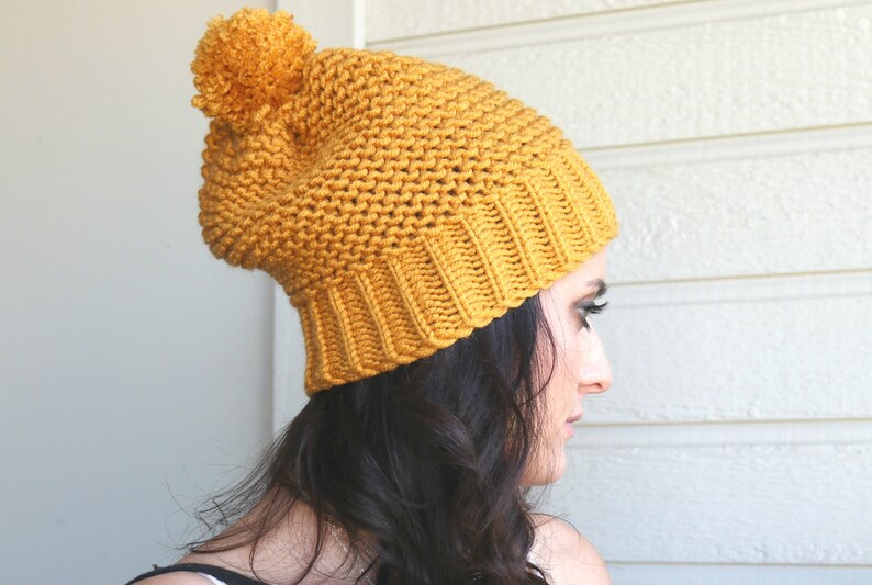 May include: A mustard yellow knitted beanie with a large pom pom on top. The beanie is slouchy and has a ribbed brim.