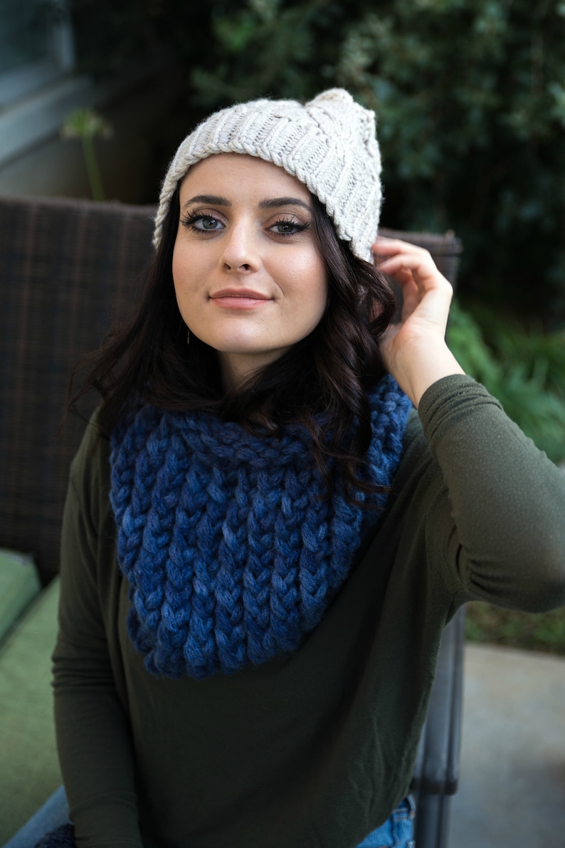 May include: A woman wearing a white knit beanie and a blue knitted scarf. She is wearing a green long-sleeved shirt and is sitting on a patio.