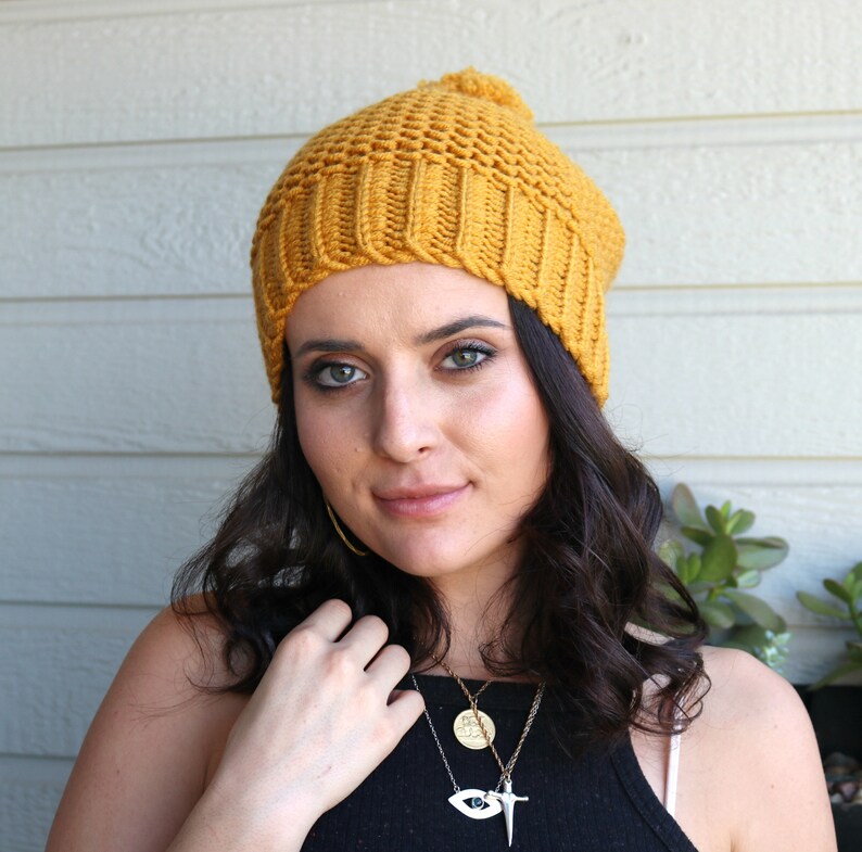 May include: A woman wearing a yellow knitted beanie with a pom pom on top. She is wearing a black tank top and several necklaces, including one with an eye pendant.