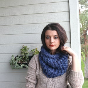 May include: A woman wearing a blue knitted infinity scarf and a beige sweater. The scarf is thick and chunky, and the sweater is a cable knit.
