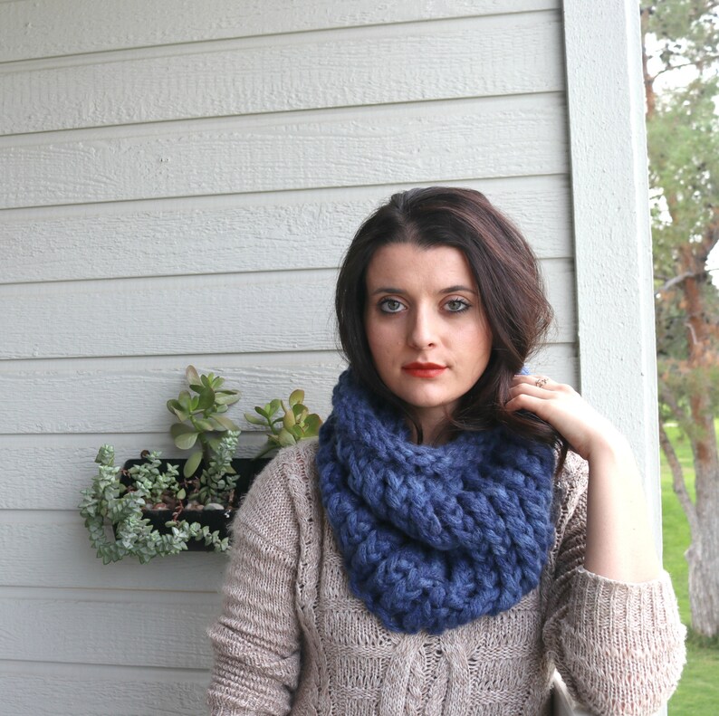 May include: A woman wearing a blue knitted infinity scarf and a beige sweater. The scarf is thick and chunky, and the sweater is a cable knit.