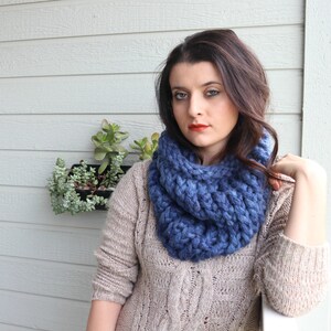 May include: A woman wearing a chunky knit blue infinity scarf with a cable knit pattern. She is wearing a beige cable knit sweater.