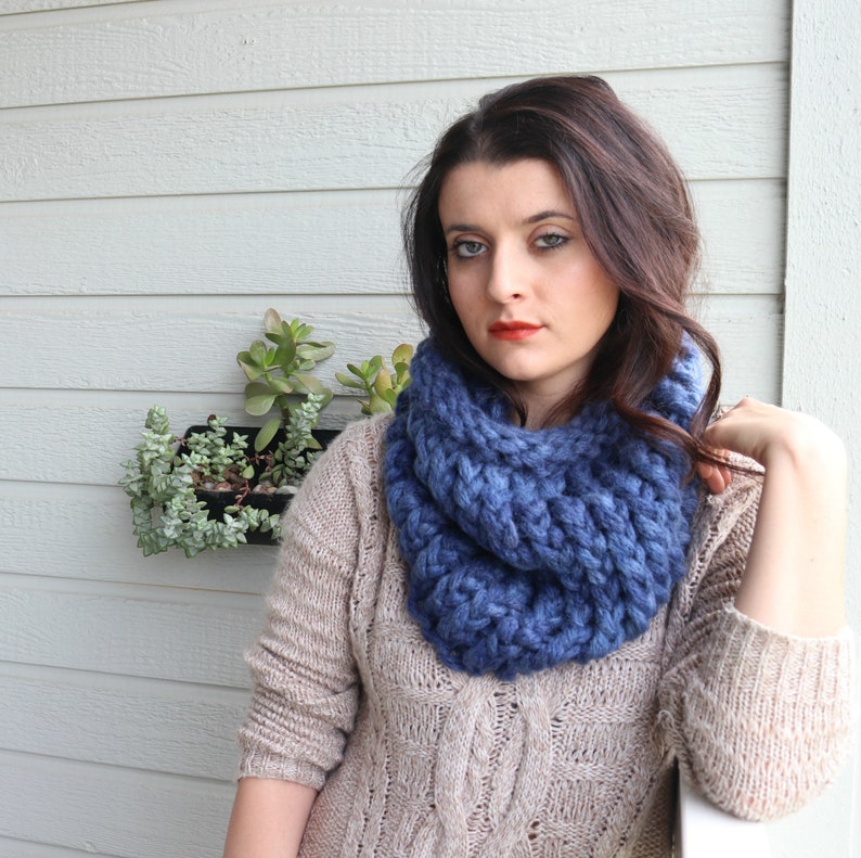 May include: A woman wearing a chunky knit blue infinity scarf with a cable knit pattern. She is wearing a beige cable knit sweater.