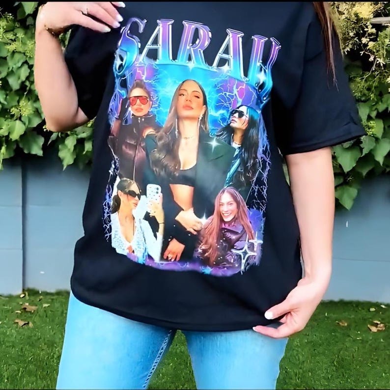 May include: Black t-shirt featuring a graphic of multiple women with the name "SARAH" in a stylized font. The graphic has a blue and purple color scheme with lightning bolt accents. The shirt is paired with blue jeans.