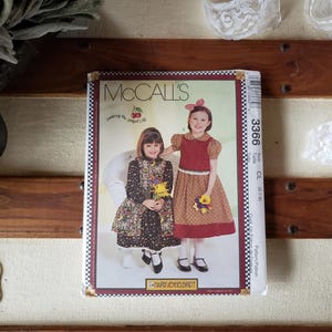May include: A vintage McCall's sewing pattern featuring two young girls in dresses. The pattern cover has a red and white checkered border and the number 3366. The background includes a wooden surface and decorative lace.