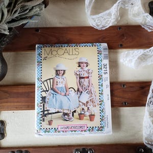 May include: A vintage McCall's sewing pattern featuring two young girls in dresses and hats. The pattern cover has a floral border and the text "McCall's". A roll of white lace and dried greenery are also visible.