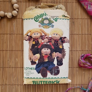 May include: A vintage Butterick pattern for Cabbage Patch Kids clothes. The pattern packet features illustrations of three Cabbage Patch Kids dolls in cowboy-themed outfits. The packet is on a bamboo mat.