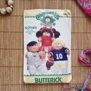 May include: A vintage Cabbage Patch Kids sewing pattern with three dolls wearing different outfits. The pattern is titled "Cabbage Patch Kids" and includes instructions for making clothes for the dolls. The pattern is from Butterick and has the number 6827.