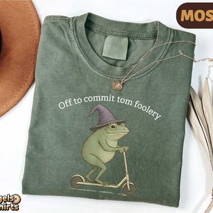 May include: A moss green t-shirt with the text "Off to commit tom foolery." The design shows a cartoon frog in a wizard hat on a scooter. A gold necklace is draped across the shirt. The t-shirt is made of cotton.