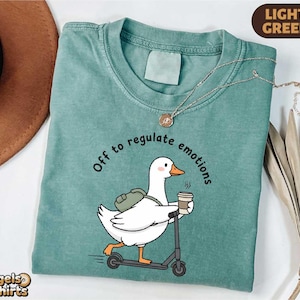 May include: Light green t-shirt with a graphic of a white duck riding a scooter and holding a coffee cup. The text "Off to regulate emotions" is printed above the duck. A gold necklace is draped across the shirt.