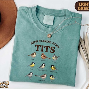 May include: A light green t-shirt with the text "STOP STARING AT MY TITS" and illustrations of various birds. The shirt has a crew neck and short sleeves. A gold necklace is draped across the shirt.
