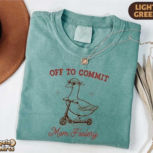 May include: Light green t-shirt with the text "OFF TO COMMIT" above a cartoon goose riding a scooter, and "Mom Foolery" below. The shirt has a crew neck and short sleeves. A gold necklace is draped across the shirt.