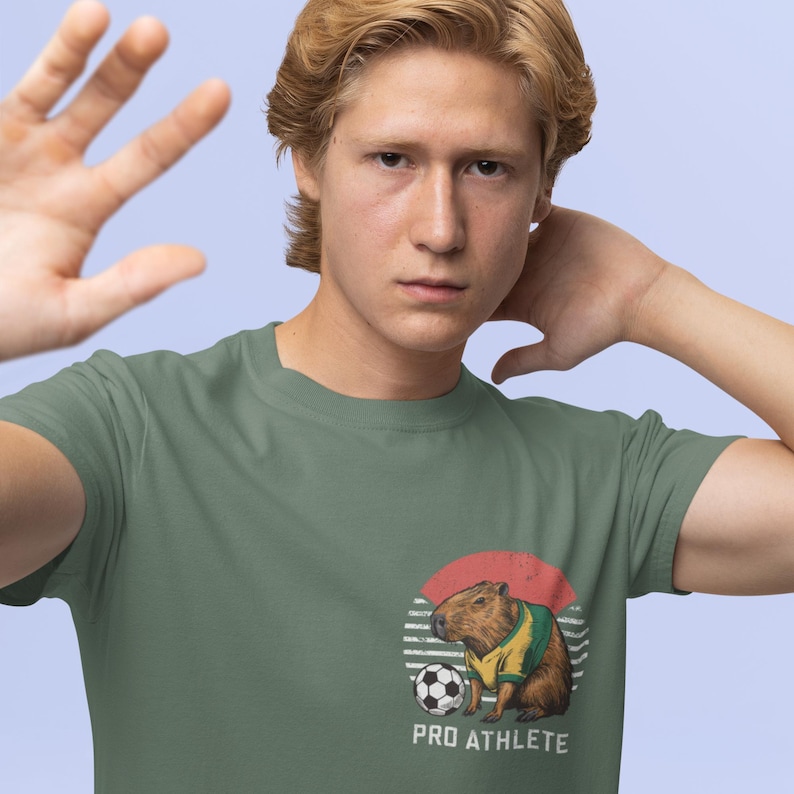 Puede incluir: Una camiseta verde con un gr&aacute;fico de un capibara vistiendo una camiseta de f&uacute;tbol y pateando un bal&oacute;n de f&uacute;tbol. El gr&aacute;fico est&aacute; rodeado por un c&iacute;rculo rojo y blanco a rayas. El texto "PRO ATHLETE" est&aacute; debajo del gr&aacute;fico.