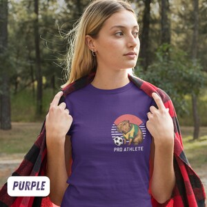 Puede incluir: Camiseta morada con un gr&aacute;fico de un capibara con una camiseta de f&uacute;tbol y sosteniendo un bal&oacute;n de f&uacute;tbol. El gr&aacute;fico est&aacute; sobre un fondo de puesta de sol retro. El texto "PRO ATHLETE" est&aacute; impreso debajo del gr&aacute;fico.