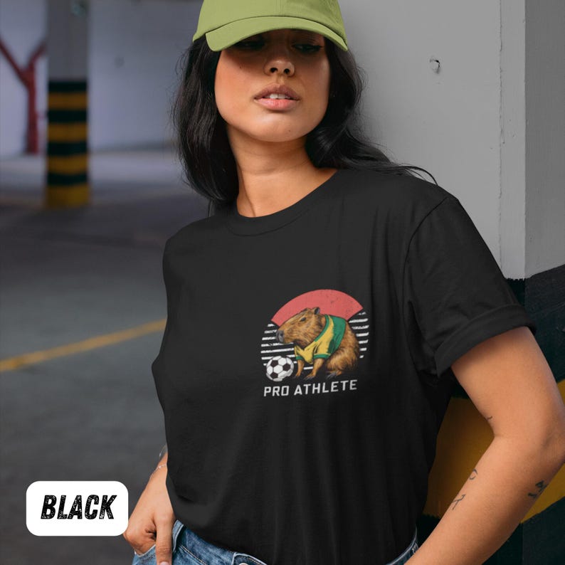 Puede incluir: Camiseta negra con un gr&aacute;fico de un capibara vistiendo una camiseta de f&uacute;tbol y pateando un bal&oacute;n de f&uacute;tbol. El gr&aacute;fico est&aacute; en un c&iacute;rculo con un fondo rojo y blanco. El texto "PRO ATHLETE" est&aacute; debajo del gr&aacute;fico.
