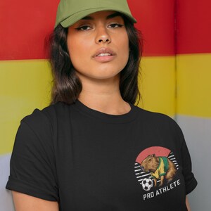 Puede incluir: Una camiseta negra con un gr&aacute;fico de un capibara vistiendo una camiseta de f&uacute;tbol y sosteniendo un bal&oacute;n de f&uacute;tbol. El gr&aacute;fico est&aacute; en un c&iacute;rculo con el texto "PRO ATHLETE" debajo.