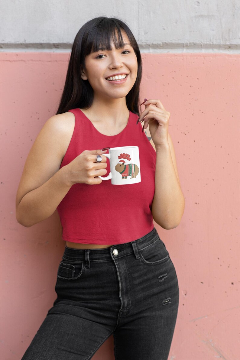 Pu&ograve; includere: Tazza in ceramica bianca con un capibara dei cartoni animati con cappello e maglione di Babbo Natale. La tazza ha la scritta "Merry" sopra il capibara. La persona indossa una canotta rossa e jeans neri.