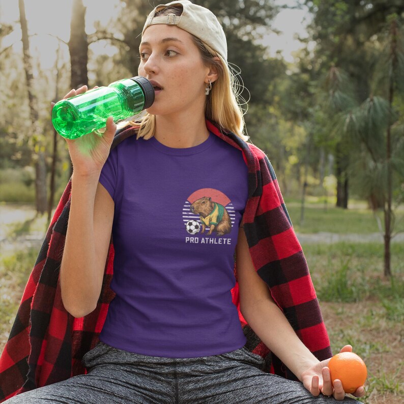 Puede incluir: Camiseta morada con un gr&aacute;fico de un capibara vistiendo una camiseta de f&uacute;tbol y sosteniendo un bal&oacute;n de f&uacute;tbol. El gr&aacute;fico est&aacute; sobre un fondo de puesta de sol con el texto "PRO ATHLETE" debajo.
