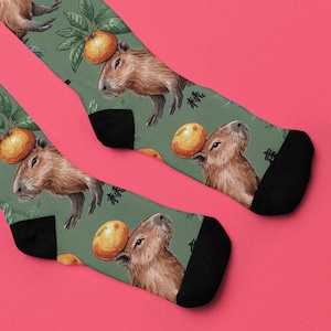 May include: A pair of green socks featuring a repeating pattern of capybaras balancing oranges on their heads. The socks have black heels and toes, with Japanese characters printed on the side. The background is a muted green.
