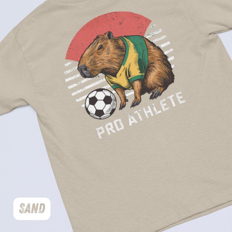 Puede incluir: Una camiseta beige con un gr&aacute;fico de una capibara vistiendo una camiseta de f&uacute;tbol y pateando un bal&oacute;n de f&uacute;tbol. El gr&aacute;fico est&aacute; en un estilo retro con un fondo a rayas rojo y blanco. El texto "PRO ATHLETE" est&aacute; debajo del gr&aacute;fico.
