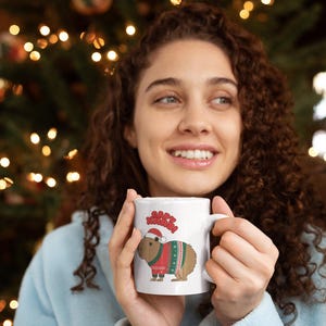Pu&ograve; includere: Tazza in ceramica bianca con un design festivo. La tazza presenta un capibara con un cappello da Babbo Natale e un maglione natalizio, con le parole "Happy Xmas!" sopra. La tazza &egrave; tenuta da una persona.