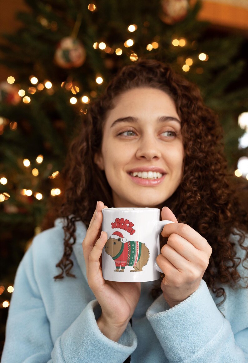 Pu&ograve; includere: Tazza in ceramica bianca con un design festivo. La tazza presenta un capibara con un cappello da Babbo Natale e un maglione natalizio, con le parole "Happy Xmas!" sopra. La tazza &egrave; tenuta da una persona.