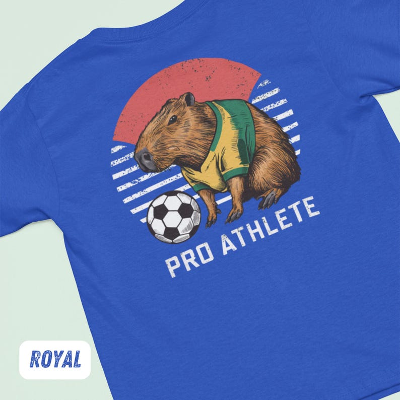 Puede incluir: Una camiseta azul real con un gr&aacute;fico de una capibara vistiendo una camiseta de f&uacute;tbol y pateando un bal&oacute;n de f&uacute;tbol. El gr&aacute;fico est&aacute; sobre un fondo rayado rojo, blanco y azul con el texto "PRO ATHLETE" debajo del gr&aacute;fico.