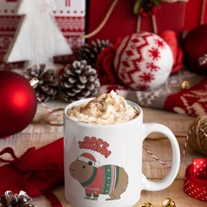 Pu&ograve; includere: Una tazza di ceramica bianca con manico, piena di una bevanda guarnita con panna montata e cannella. La tazza presenta un capibara con cappello e maglione di Babbo Natale, con la scritta "Happy Xmas!". Decorazioni natalizie.