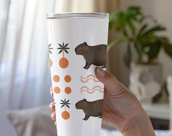 Può includere: Un bicchiere bianco con un coperchio che presenta un capibara, palme e cerchi arancioni. Il capibara è marrone e ha la pancia bianca. Le palme sono verdi e hanno cerchi arancioni. Il bicchiere è tenuto da una mano.