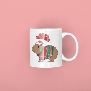 Pu&ograve; includere: Tazza in ceramica bianca con un capibara dei cartoni animati che indossa un cappello da Babbo Natale e un maglione festivo con motivi rossi, verdi e bianchi. La tazza ha le parole "Happy Xmas" sopra il capibara. Lo sfondo &egrave; rosa.
