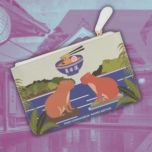 May include: A beige zippered pouch with a colorful illustration of two capybaras enjoying ramen noodles. The pouch has the text "Food Connoisseur - Ramen Edition" printed on the bottom.