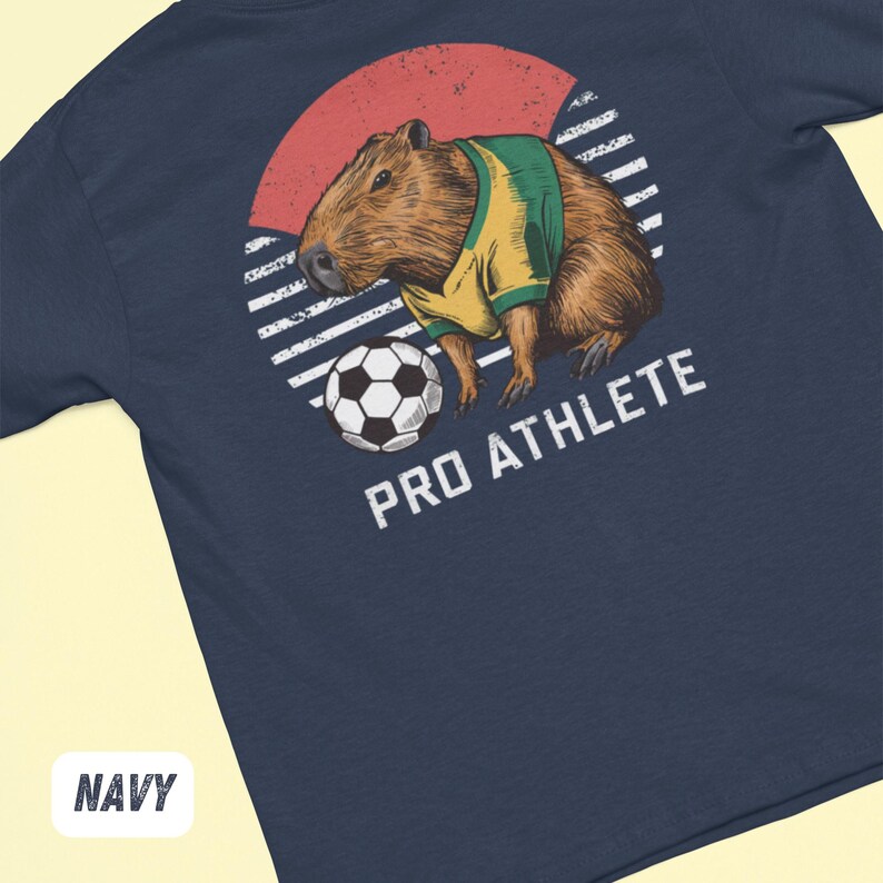 Puede incluir: Una camiseta azul marino con un gr&aacute;fico de una capibara vistiendo una camiseta de f&uacute;tbol y sosteniendo un bal&oacute;n de f&uacute;tbol. El gr&aacute;fico est&aacute; sobre un fondo rayado rojo, blanco y amarillo con el texto "PRO ATHLETE" debajo del gr&aacute;fico.